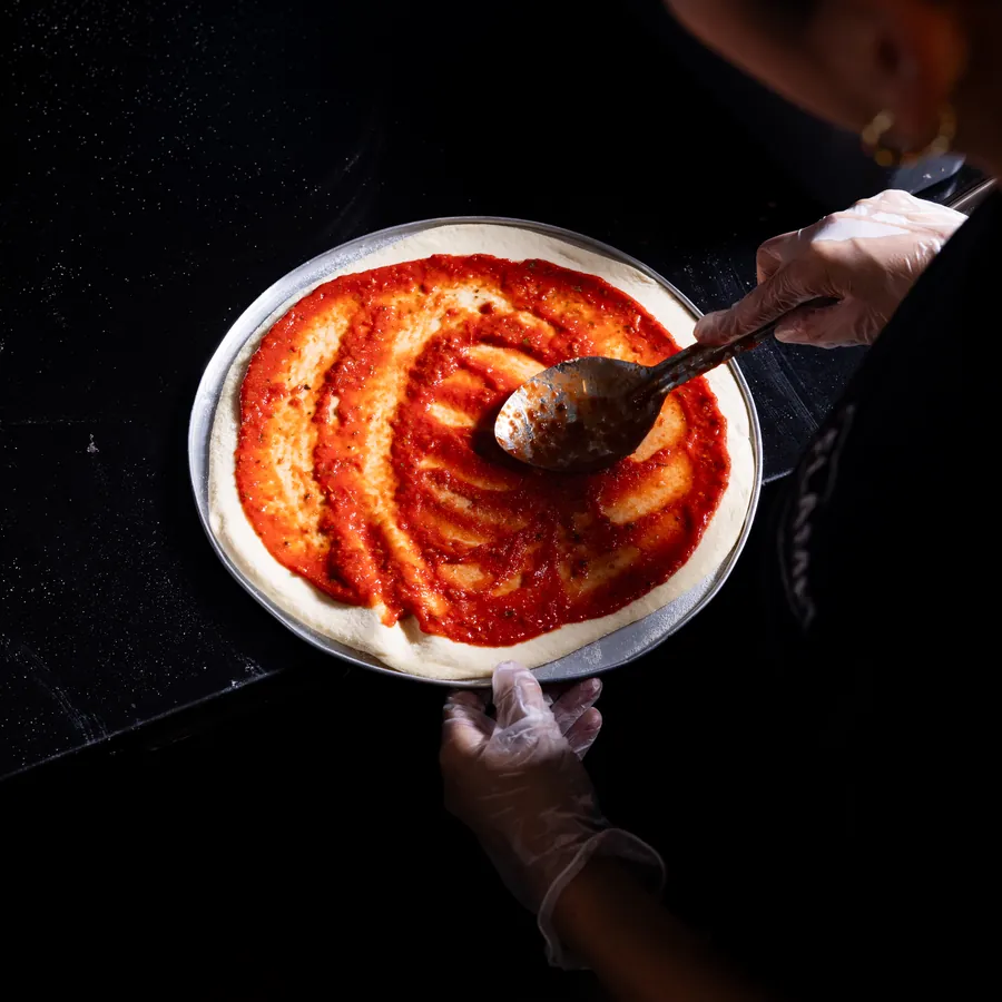 Tomato sauce being spread over a pizza base with the back of a spoon