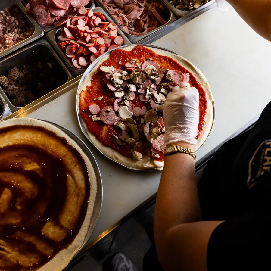 A gloved hand adding mushrooms and salami to a pizza on the bench