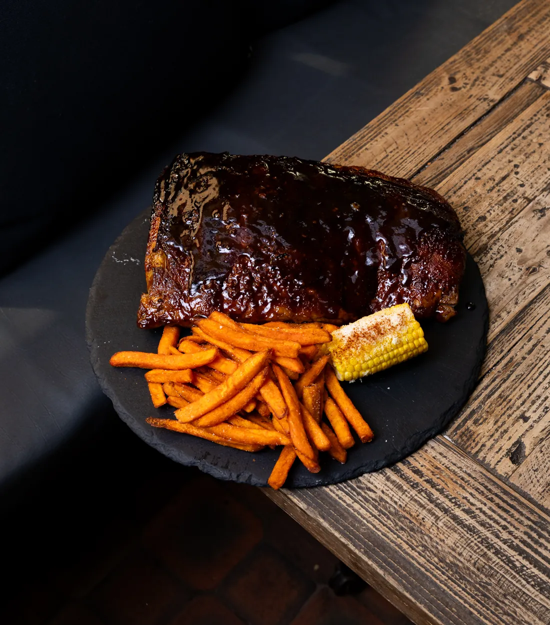 Glazed ribs with sweet potato fries and a piece of corn on a slate board