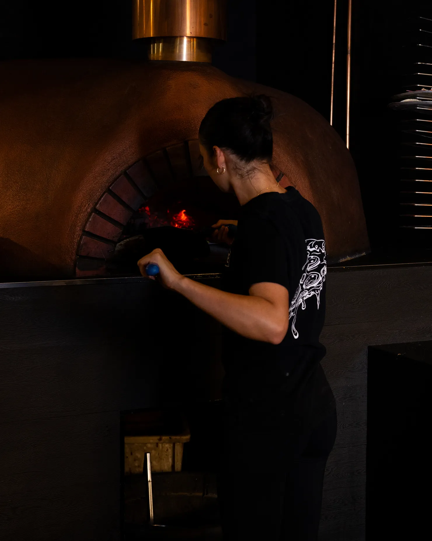 A cook tending the copper-domed woodfire oven in a dark kitchen