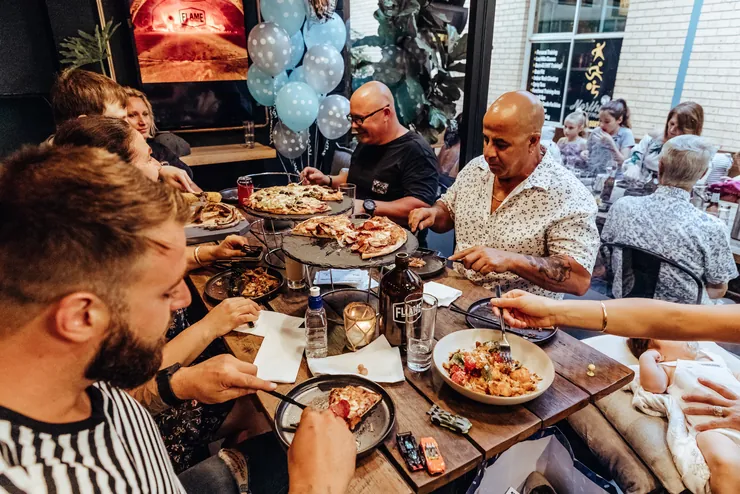 A group celebrating around a table of shared pizzas, with balloons behind them