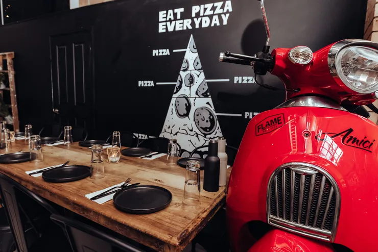 Long tables laid with black plates and glasses beside a red Vespa and a chalk pizza mural