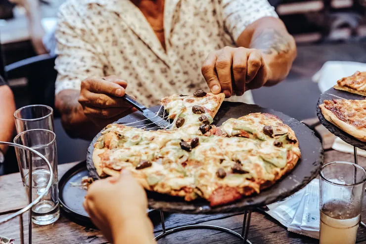 Hands reaching across the table for slices of a shared pizza on a raised stand