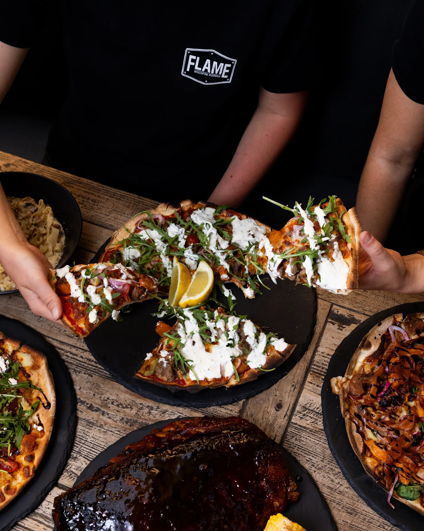Hands reaching for slices of pizza across a table crowded with shared dishes