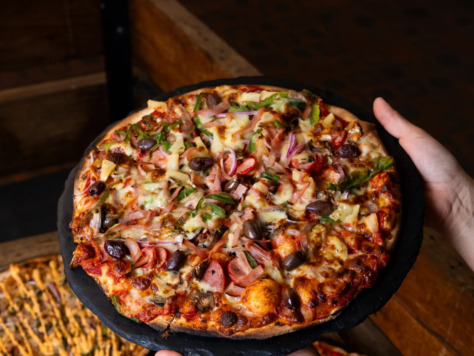 A supreme pizza on a slate board being set down on a timber table