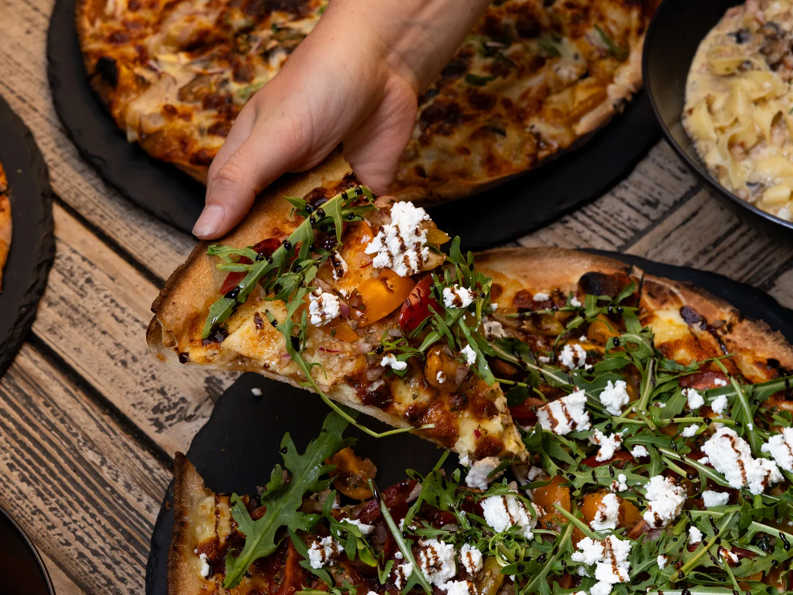 A hand lifting a slice of pizza topped with rocket and feta from a shared table