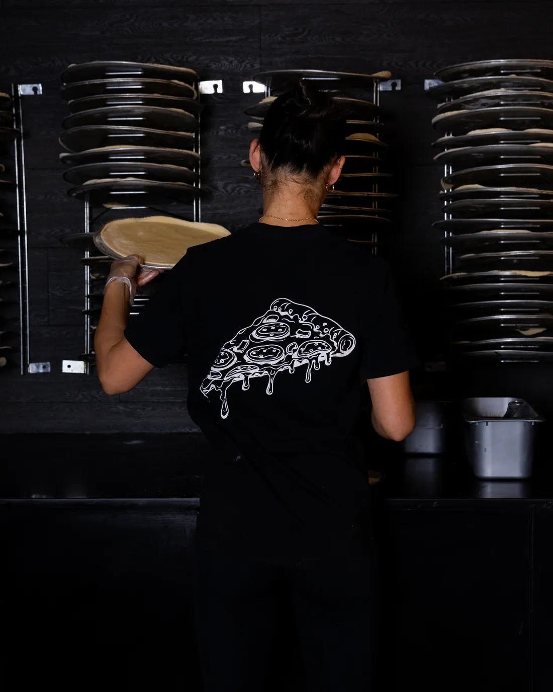 A cook in a Flame shirt stretching a pizza base, stacks of pizza trays either side