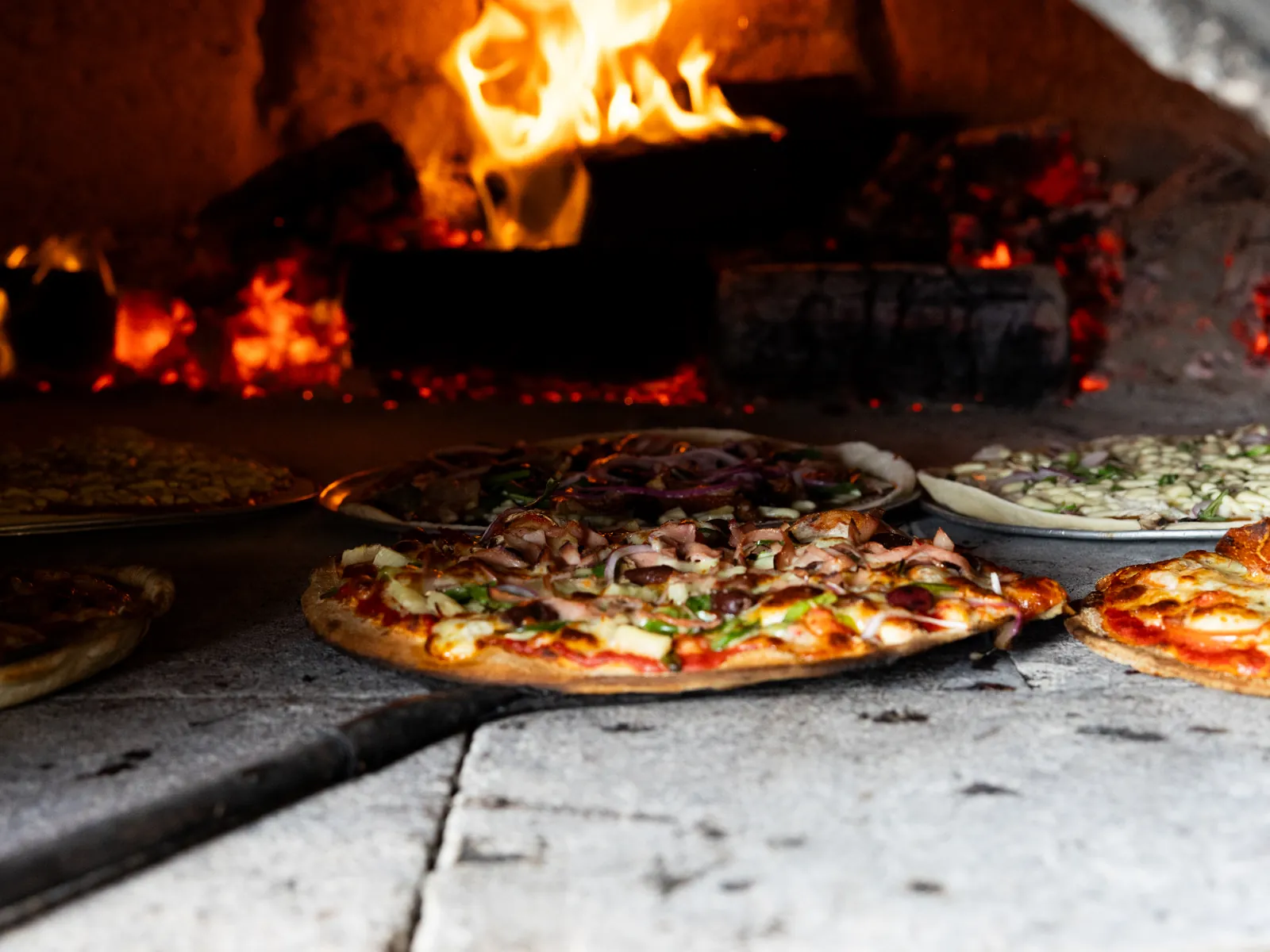 Pizzas cooking on the floor of the woodfire oven, flames behind them