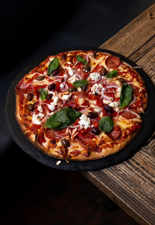 A woodfired pizza with salami, olives, feta, red onion and fresh basil on a slate board